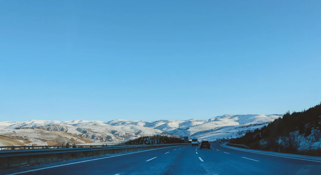 Open highway with guardrails, snow-covered mountains in the distance, under a clear blue sky.