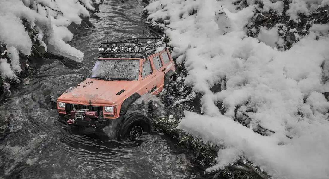 Orange off-road vehicle stuck in a snowy stream, surrounded by snow-covered rocks and trees.