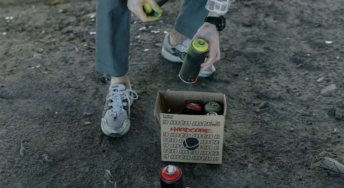Person kneeling on gravel, holding spray paint cans over a box of spray cans outdoors, ready for paint blending.
