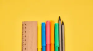 Assorted colored markers and a wooden ruler on a bright yellow background, illustrating color matching and measurement steps for car touch-up with paint pens.