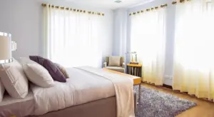 Bright bedroom interior with a neatly made bed, large windows with sheer white curtains, and a gray area rug.