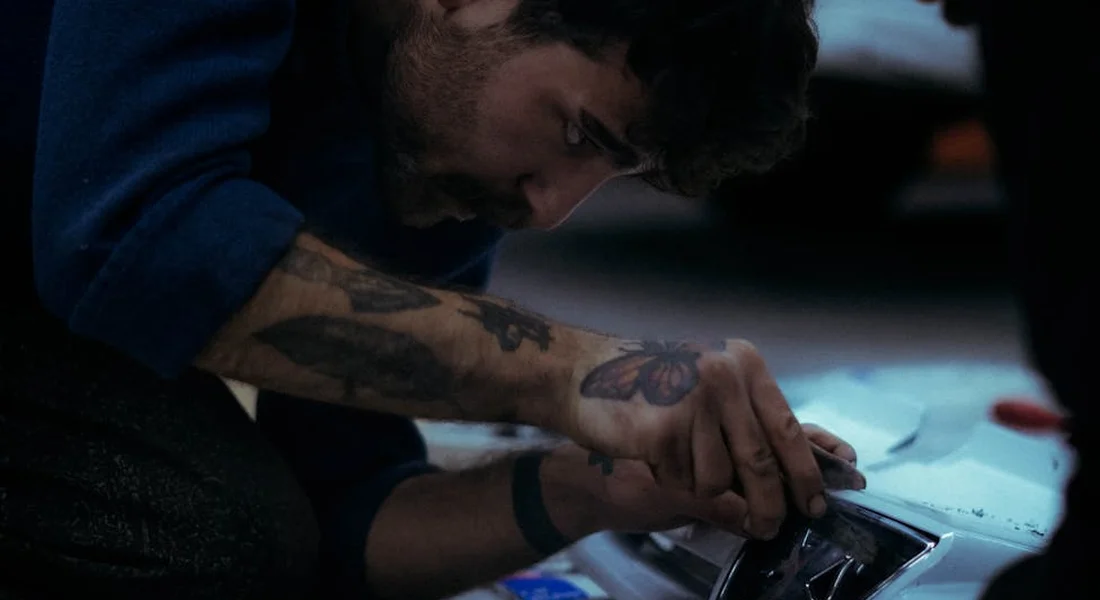 Tattooed person leaning over a car bumper while applying touch-up work