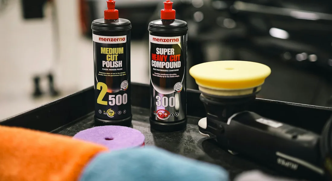 Two Menzerna car polish bottles on a detailing tray with colorful applicator pads and a car in the background.