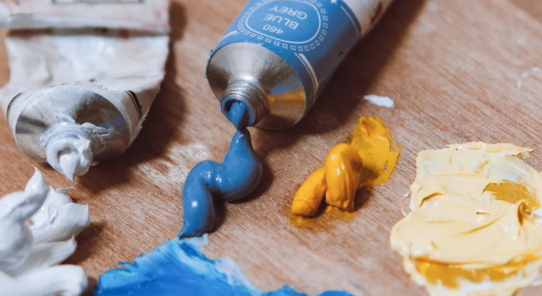 Close-up of paint tubes and blobs of blue, white, and yellow paint on a wooden workbench.