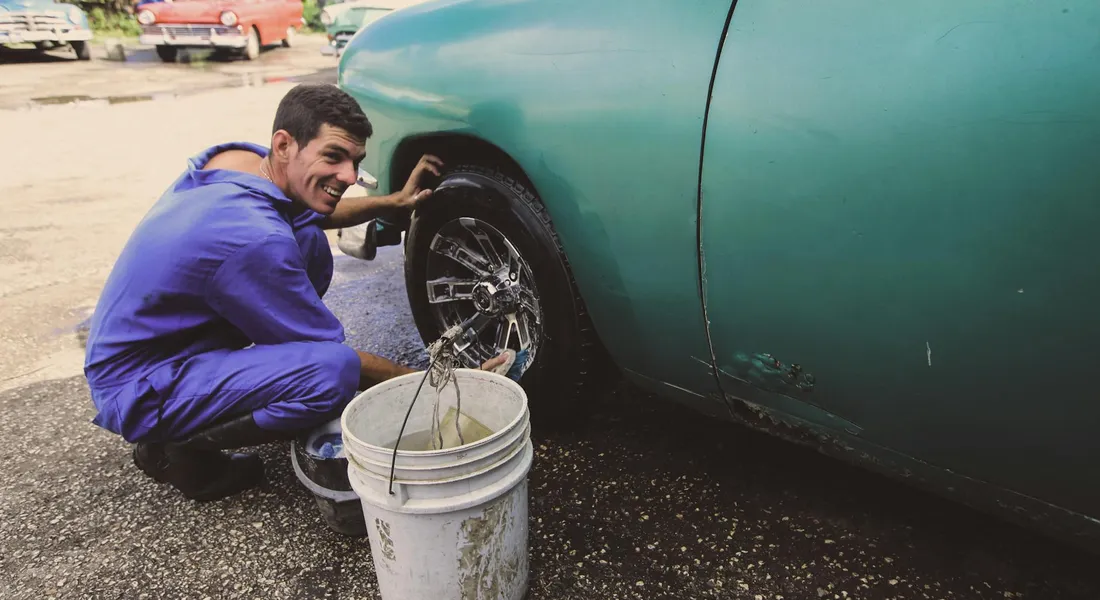 A man in a blue shirt crouches next to a green car, washing its wheel with a hose and a white bucket of soapy water.