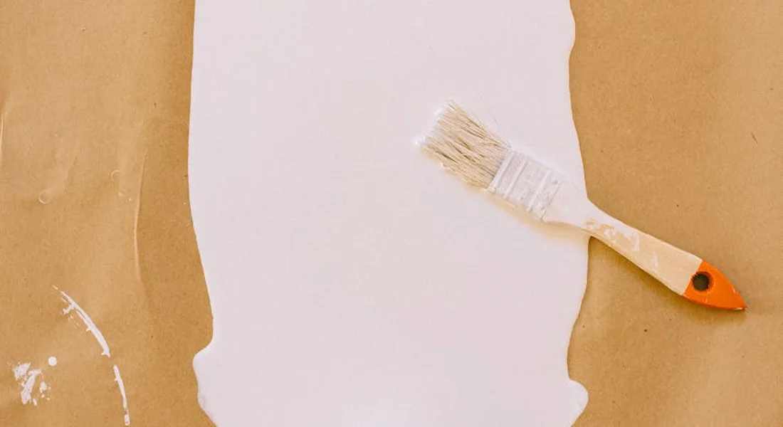 A paintbrush rests on torn white paper on a brown background, symbolizing preparation before removing spray paint.