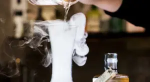 A gloved hand pours a liquid into a beaker, with car detailing products visible in the background, illustrating preparation for paint decontamination.