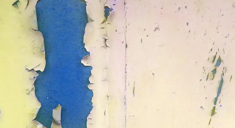 Close-up of peeling wall paint in blue and pale yellow, showing layers flaking away