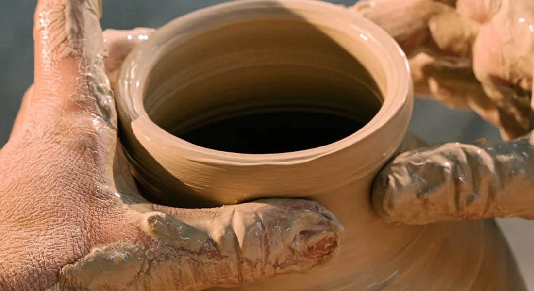 Hands covered in clay shaping a clay pot on a pottery wheel.