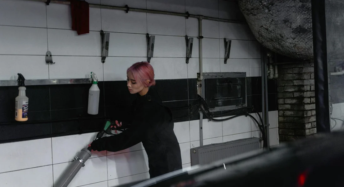 Professional car detailer with pink hair wearing black clothing uses a cleaning tool inside a tiled workshop, with car detailing equipment on the wall.
