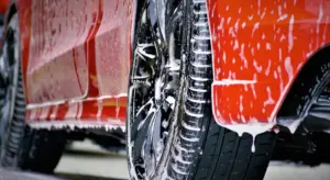 Close-up of a red car being washed, showing soap on the exterior and a shiny wheel to illustrate exterior detailing services.