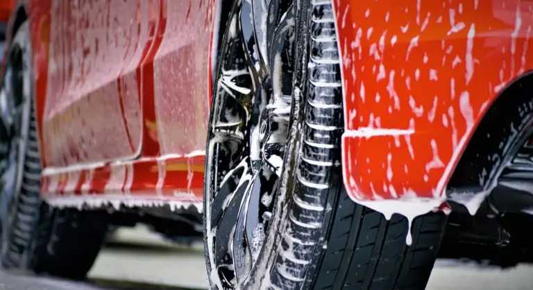 Close-up of a red car being washed, showing soap on the exterior and a shiny wheel to illustrate exterior detailing services.