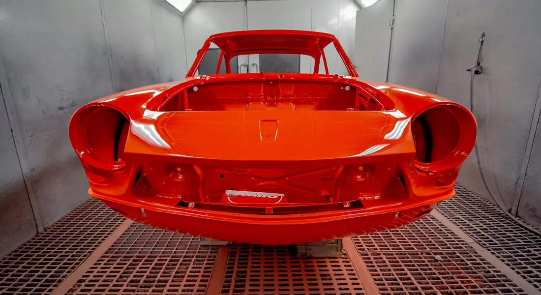 Red car body shell inside a professional paint booth, ready for restoration.