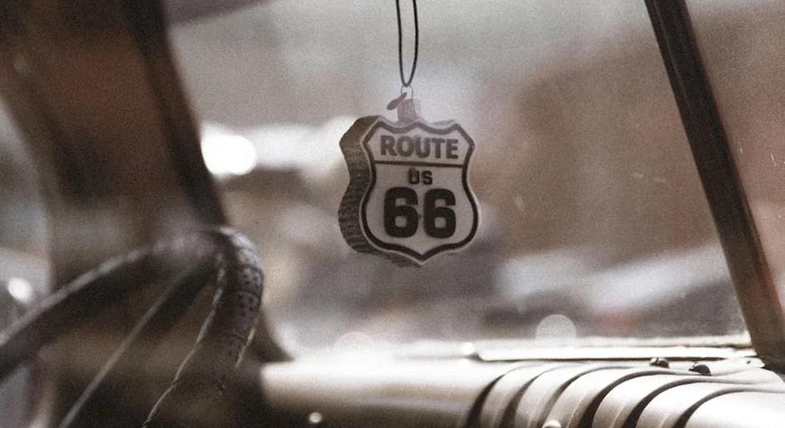 Route 66 shield pendant hanging from a car's rearview mirror inside a vintage sedan, with a blurred dashboard in the background.