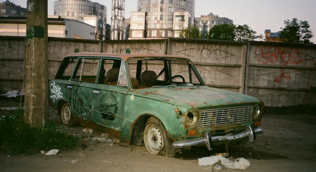 Old green car with rust and paint flaking in a scrapyard, surrounded by graffiti-covered walls