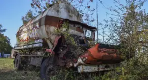 Extensively rusted and damaged vehicle with overgrown vegetation around the body and exposed interior components.