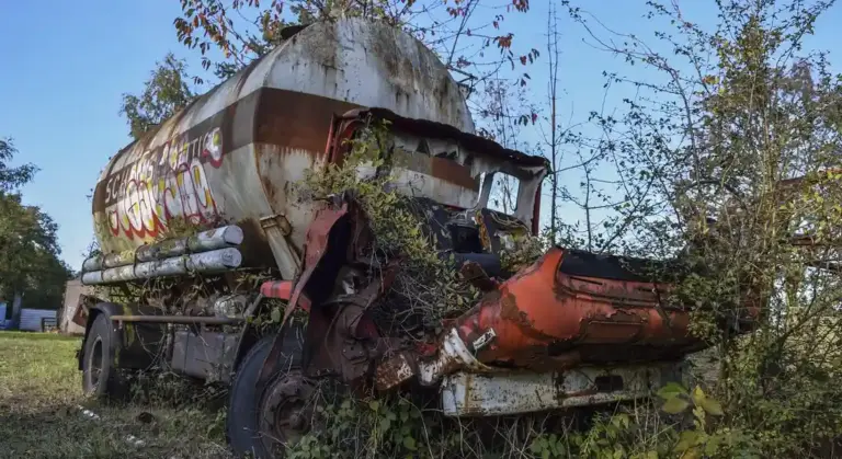 Extensively rusted and damaged vehicle with overgrown vegetation around the body and exposed interior components.