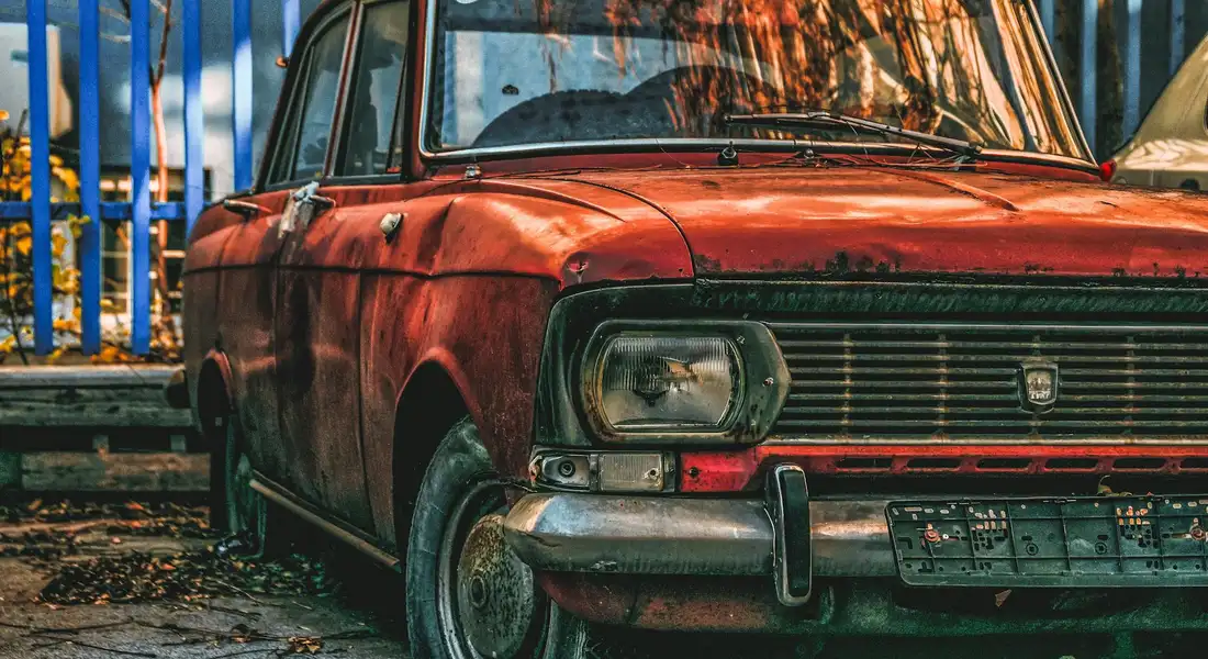 Old red car with rust on the body panels and a worn bumper, parked outdoors.