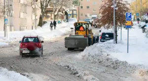 Red SUV driving on a snow-covered street with a snowplow clearing the road; snowbanks line the sides and other vehicles are parked along the road.