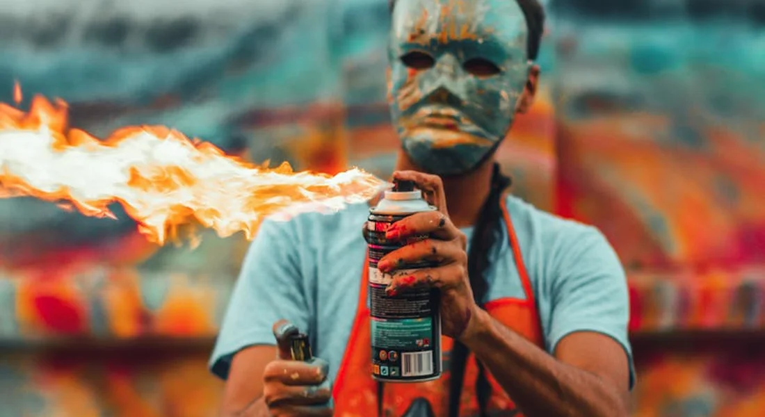 Person wearing a protective mask and apron holds a spray can in front of a colorful graffiti wall; a flame from a lighter is visible near the can.