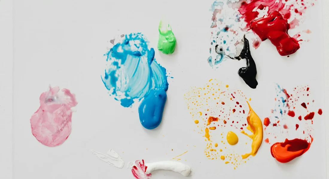 Close-up of multicolored spray paint blobs and splatter on a white surface, representing dried-on paint on a vehicle exterior.