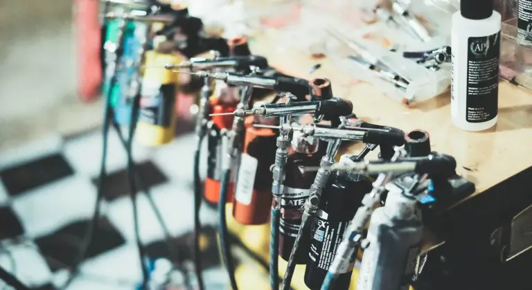 Row of spray guns hanging on a rack above a wooden workbench, with a bottle of solvent visible in the background.