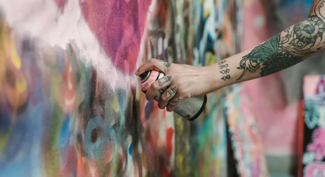 Tattooed arm holding a spray paint can spraying colorful graffiti on a mural