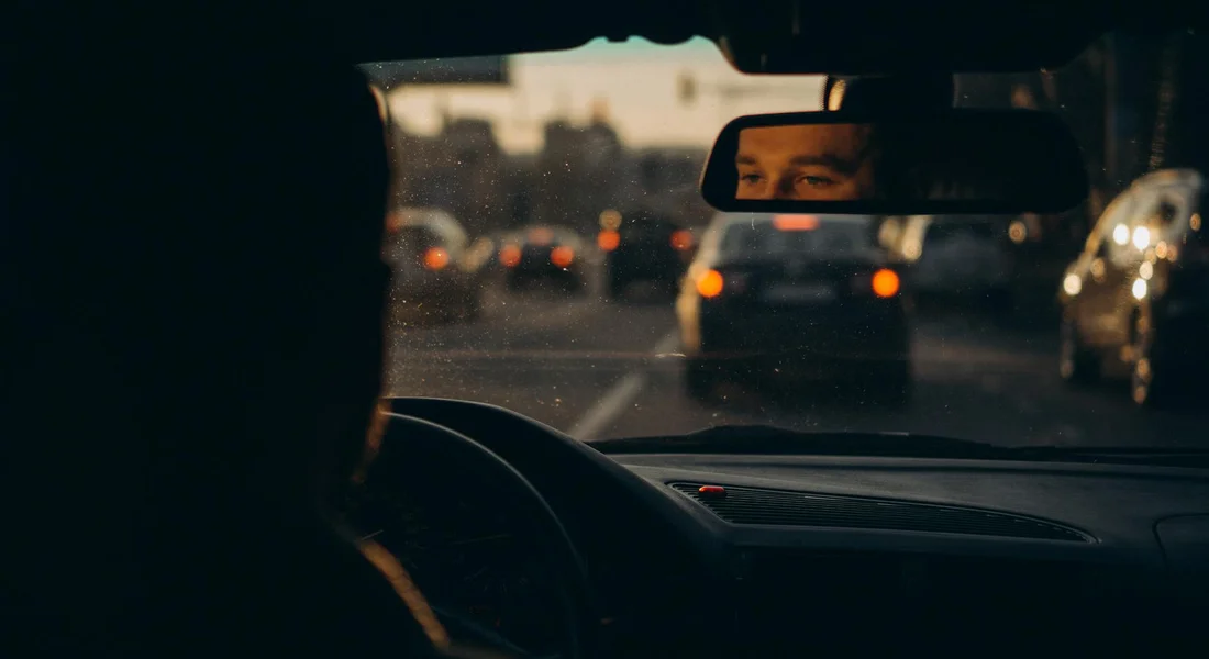 From the back seat, a driver is seen inside a car with traffic visible through the windshield, illustrating the car interior in a dim, dusk-lit setting.