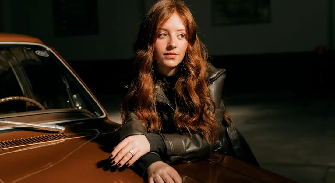 Young woman with long red hair wearing a dark jacket leaning on a vintage orange car