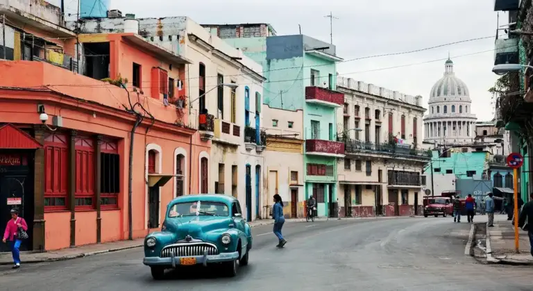 A vintage blue car drives down a colorful street lined with pastel buildings; a domed building is visible in the distance.