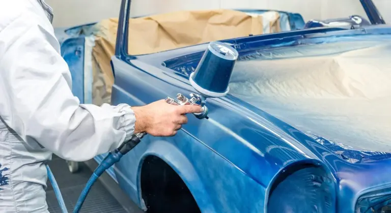 Technician wearing a protective suit spray-paints blue car in a paint booth to repair sun damage and fading.