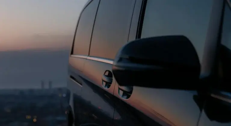 Close-up of a car’s tinted side window and mirror with a sunset reflection.