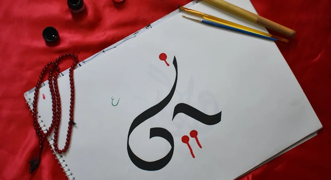 A white sketchbook with a bold black calligraphic symbol, surrounded by red beads and pencils on a red fabric background.