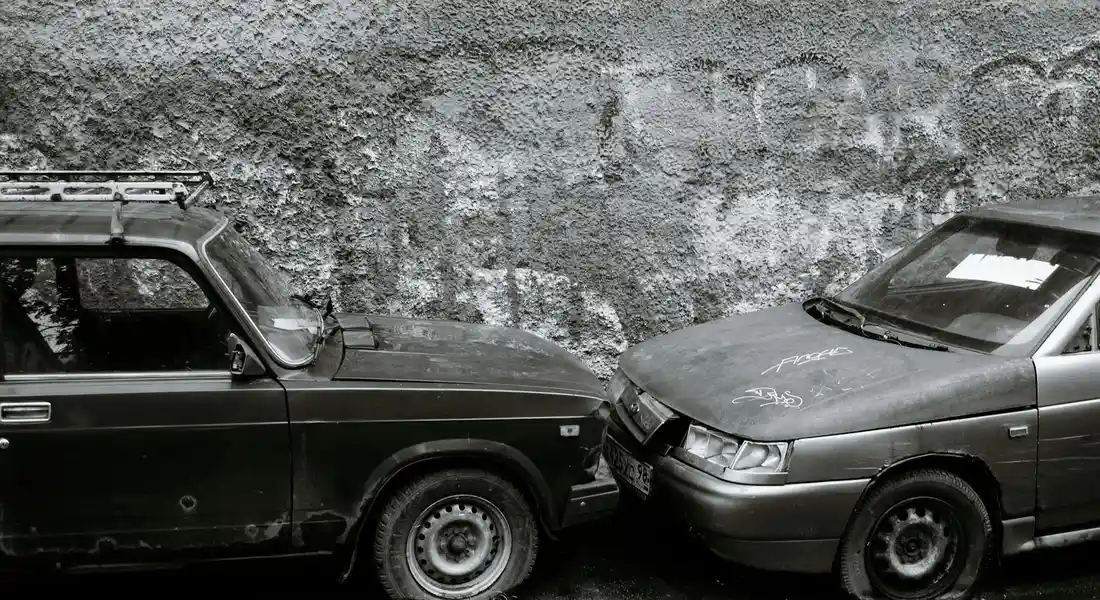 Two cars with visible collision damage and scratches parked in front of a textured wall.