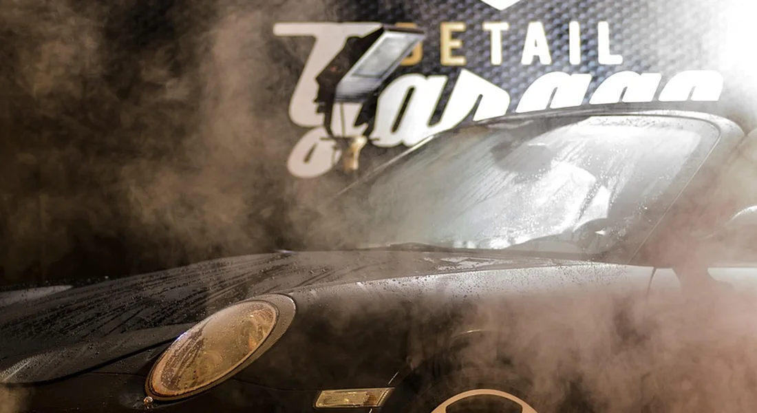 A dark car being washed with mist from a spray, with detailing signage visible in the background.