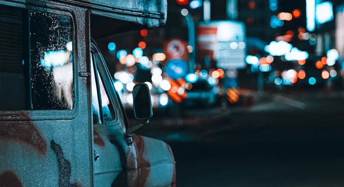 Side view of a weathered van with bug splatters on its door and window, parked on a city street at night with blurred neon lights.