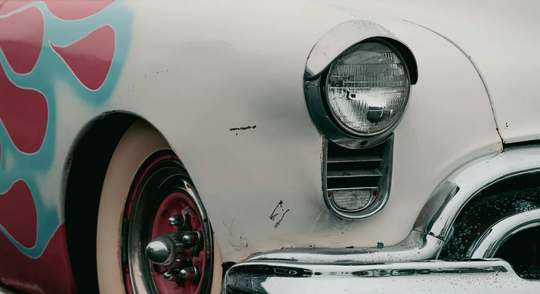 Close-up of a vintage car's front quarter panel with a turquoise and pink mural on the left, a round headlight, chrome bumper, and visible paint scratches.
