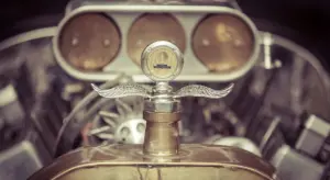 Close-up of a vintage car hood ornament with an exposed engine in the background, emphasizing the vehicle's glossy exterior finish.