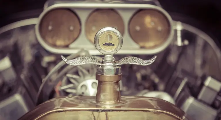 Close-up of a vintage car hood ornament with an exposed engine in the background, emphasizing the vehicle's glossy exterior finish.