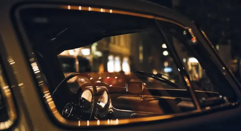 Close-up view of a vintage car's dashboard and steering wheel through a window with reflections at night