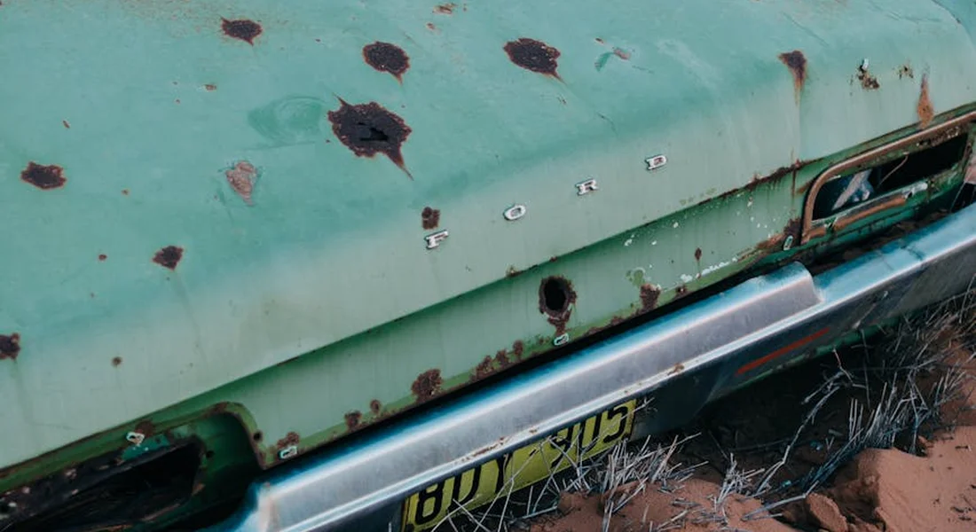 Close-up of a vintage green car with rust holes and a chrome bumper, resting on dirt.