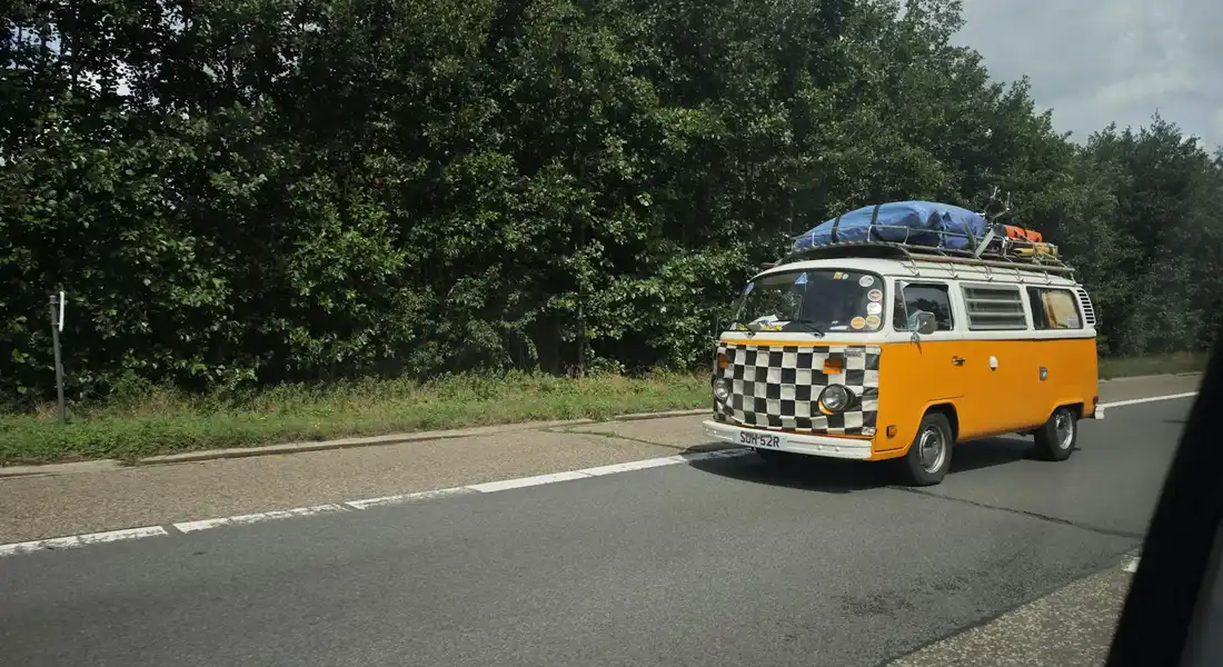 Orange and white vintage van with a checkered front, roof rack loaded with luggage, driving on a highway with trees lining the road.