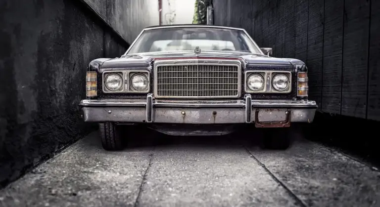 Front view of a vintage car parked in a narrow, shadowy alley with a chrome grille and weathered bumper.