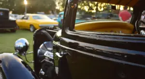 Close-up of a glossy black vintage car door with chrome trim, blurred yellow and other cars in the background.