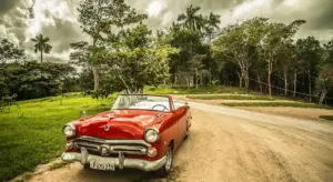 Red vintage convertible on a dirt road surrounded by green trees under a cloudy sky.