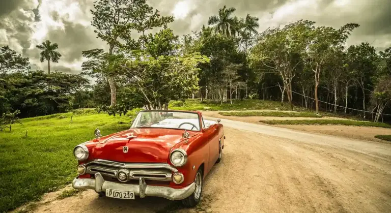 Red vintage convertible on a dirt road surrounded by green trees under a cloudy sky.