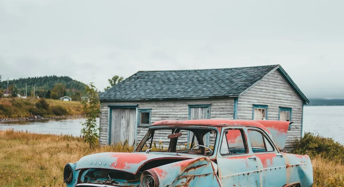 A faded blue and red vintage car with rust patches sits beside a lakeside shack in a grassy field.