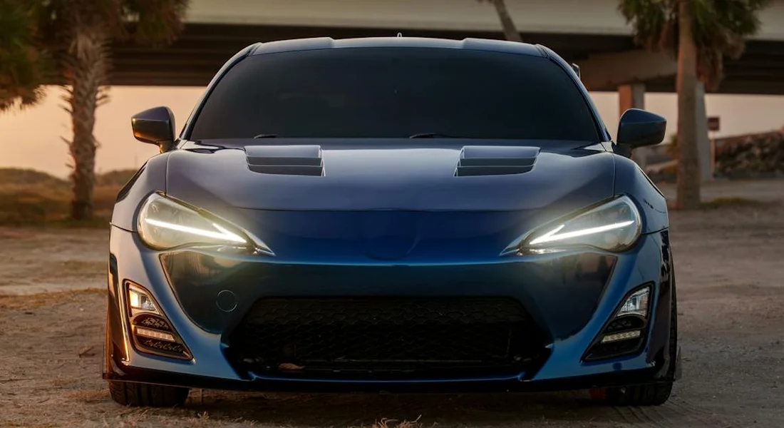 Front view of a blue vinyl-wrapped sports car with headlights on, parked outdoors at sunset