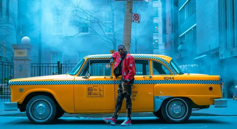 A man in a red jacket stands beside a bright yellow taxi with blue smoke in the background on an urban street.