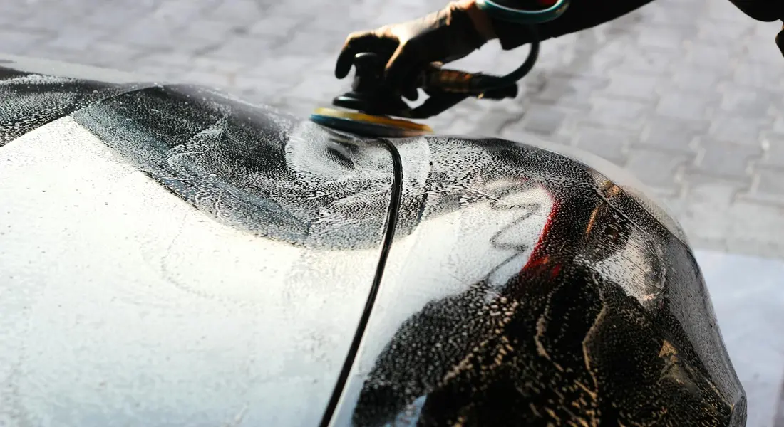 Close-up of a car's glossy hood being buffed with a polishing tool, with droplets of water and wax on the surface.